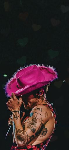 a tattooed man wearing a pink hat and holding his hands up to his face while standing in front of a black background