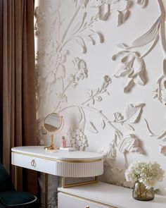 a white dresser with a mirror and flowers on it next to a wallpapered room