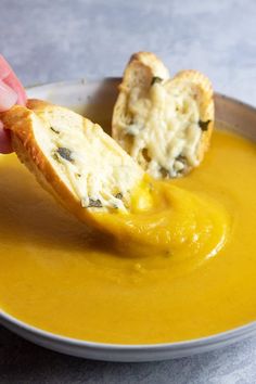 a piece of bread is being dipped into a bowl of yellow soup with cheese on top