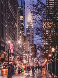 people are walking down the street at night in new york city, with skyscrapers lit up behind them