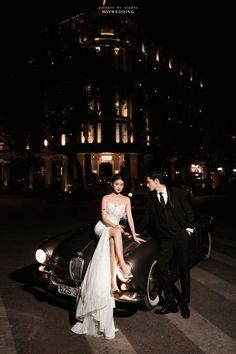 a bride and groom sitting on the hood of a vintage car in front of a building at night