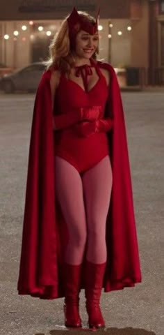 a woman in a red bodysuit and cape standing on the street with her hands behind her back