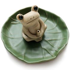 a ceramic frog sitting on top of a leaf shaped plate with eyes wide open and one eye partially closed