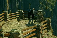 two people are standing on a wooden bench overlooking the mountains and trees in the distance