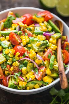 a white bowl filled with corn, avocado and tomato salad next to a wooden spoon