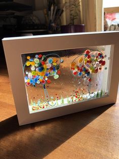 an art work is displayed in a white frame on a wooden table next to a potted plant