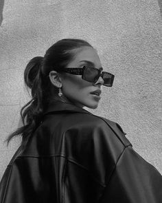 a woman with sunglasses on her face and wearing a black leather jacket, standing in front of a concrete wall