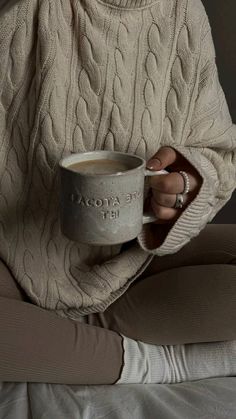 a woman sitting on a bed holding a cup of coffee in her hands and wearing a sweater