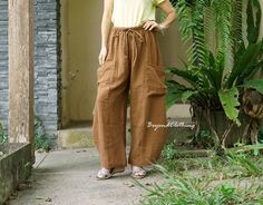 a woman standing in front of some plants wearing brown pants and a yellow t - shirt