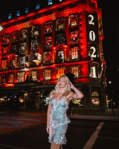 a woman standing on the street in front of a building with lights all over it