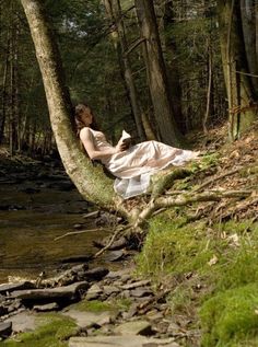 a woman in a white dress sitting on a tree next to a river and rocks