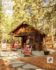 a small cabin in the woods with red chairs and fire pit on it's patio