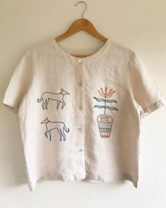 a white shirt with embroidered animals and flowers on the front, hanging from a wooden hanger