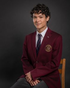 a young man in a maroon suit and tie sitting on a chair with his arms crossed