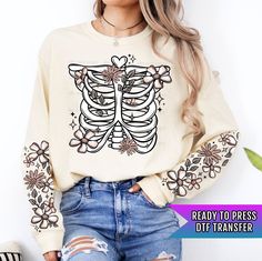 a woman wearing ripped jeans and a sweater with a skeleton print on the sleeves, standing in front of a white wall