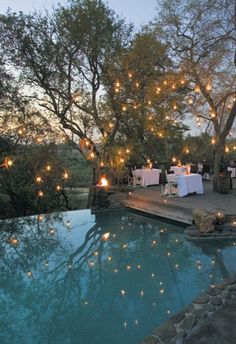 an outdoor dining area next to a swimming pool with lights strung from the trees over it