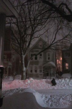 a snow covered street with houses and trees in the background at night, as seen from across the street