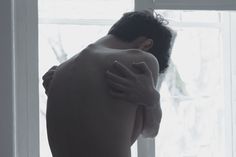 a shirtless man standing in front of a window with his arm wrapped around him