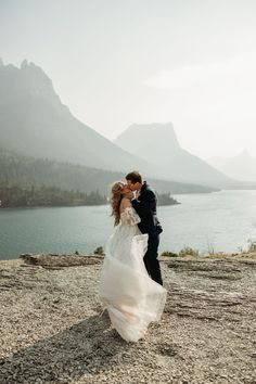 a bride and groom kissing on top of a mountain overlooking the water with mountains in the background