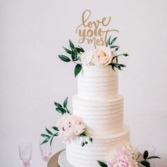 a white wedding cake with flowers and greenery on the top is decorated with gold lettering love you most