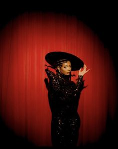 a woman in a black dress and hat posing with her hands on the side of her head