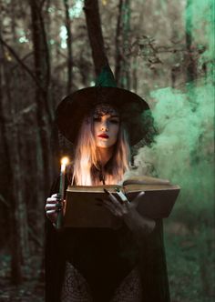 a woman dressed as a witch reading a book in the woods with a lit candle