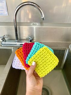 a hand is holding four colorful scrubcloths in front of a sink with a faucet