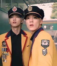 two people in uniforms standing next to each other near a train station and one person is looking at the camera