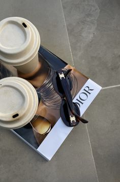 two coffee cups sitting on top of a magazine next to sunglasses and a pair of glasses