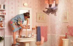 a man standing on top of a toilet next to a bathroom sink filled with lots of clutter