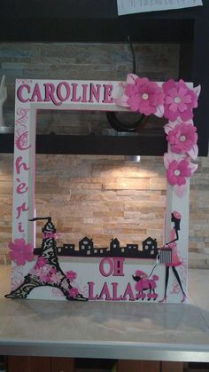 a pink and white photo frame with flowers on the top, in front of a brick wall