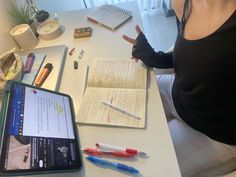 a woman sitting at a desk with an open book and notebooks on it, pointing to the screen