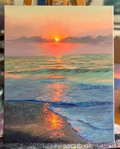 an easel with a painting on it and the sun setting over the ocean