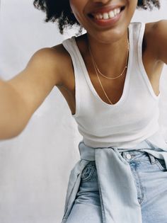 a woman wearing a white tank top and blue jeans with her arms behind her head