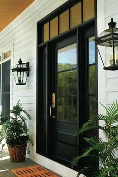 a black front door on a white house with potted plants and two lights hanging from the side