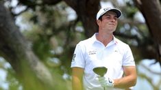 a man holding a golf club in his right hand while standing next to a tree