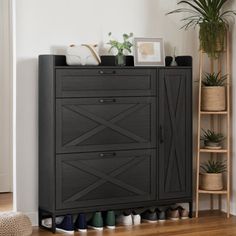 a black cabinet with shoes on it in front of a white wall and potted plant