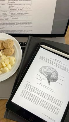 the tablet is next to a bowl of food and an open book on a desk