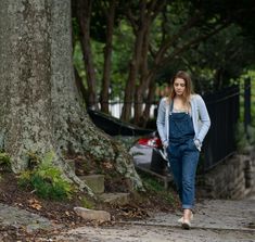 a woman in overalls walking down a sidewalk next to a large tree and fence