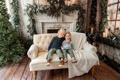 two children sitting on a couch in front of a fireplace with christmas decorations and greenery