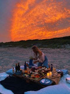 a woman sitting on the beach with food and drinks in front of an orange sky