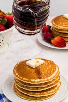 a stack of pancakes with syrup being poured on top and strawberries in the background