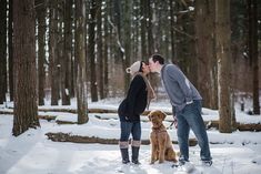 a man and woman kissing in the woods with their dog on a leash, surrounded by snow