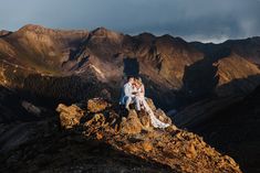 a bride and groom sitting on top of a mountain at sunset with mountains in the background