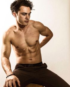 a shirtless young man sitting on top of a wooden chair next to a wall