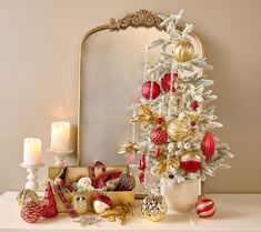 a white christmas tree with red and gold ornaments in front of a mirror, lit candles and other holiday decorations