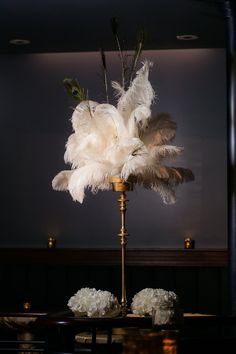 two white flowers and feathers on a gold stand in front of a dark room with black walls