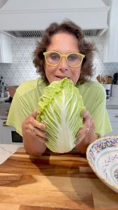 a woman in glasses is holding up a large cabbage leaf while sitting at a kitchen counter