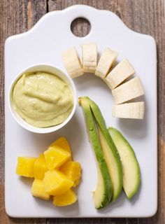 a white cutting board topped with sliced fruit and avocado next to a bowl of dip