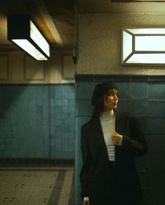 a woman standing in front of a blue tiled wall wearing a black suit and white turtle neck sweater